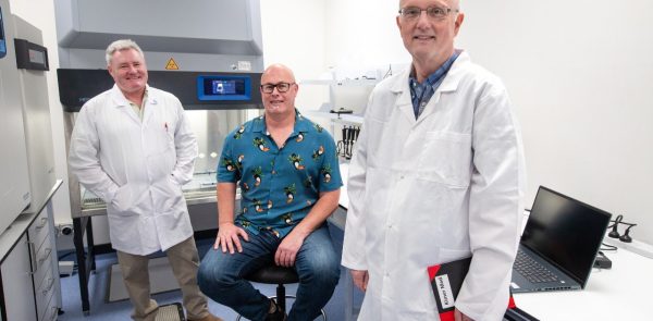 Two Men In White Lab Coats Standing In A Science Lab, While Another Man In Jeans And A Colourful Shirt Is Seated Between Them.
